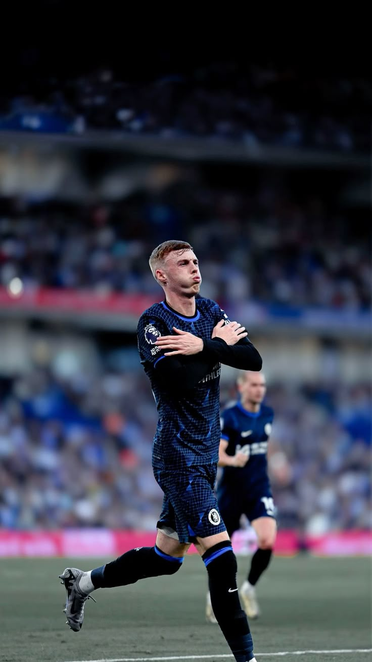 Cole Palmer of Chelsea jogging with arms crossed across his chest in the 'cold' celebration, dark blue Chelsea away kit, packed stadium behind him.