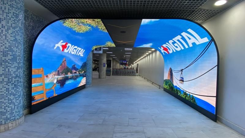 K-Digital airport corridor with two curved LED tunnel-arches built into the walls, each panel displaying Rio de Janeiro scenery: Sugarloaf Mountain, the Sao Conrado beach view, and the Sugarloaf cable car. Polished stone floor between the arches.