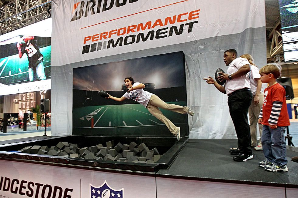 Bridgestone 'Performance Moment' NFL activation: a woman in a white tee dives onto a foam-cube pit toward a large screen showing an endzone scene, with a coach holding a football and a boy watching from a small platform on the right.